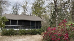 A house with a porch and large windows, surrounded by trees and a garden with pink flowers. - Wakulla River Retreat- Manatee Cottage  (Crawfordville)