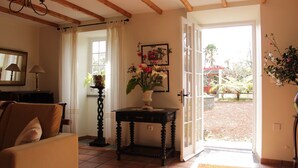 Une pièce comprenant un canapé, une table ornée d’un vase de fleurs, un miroir et une vue sur le jardin, visible à travers une porte ouverte.
