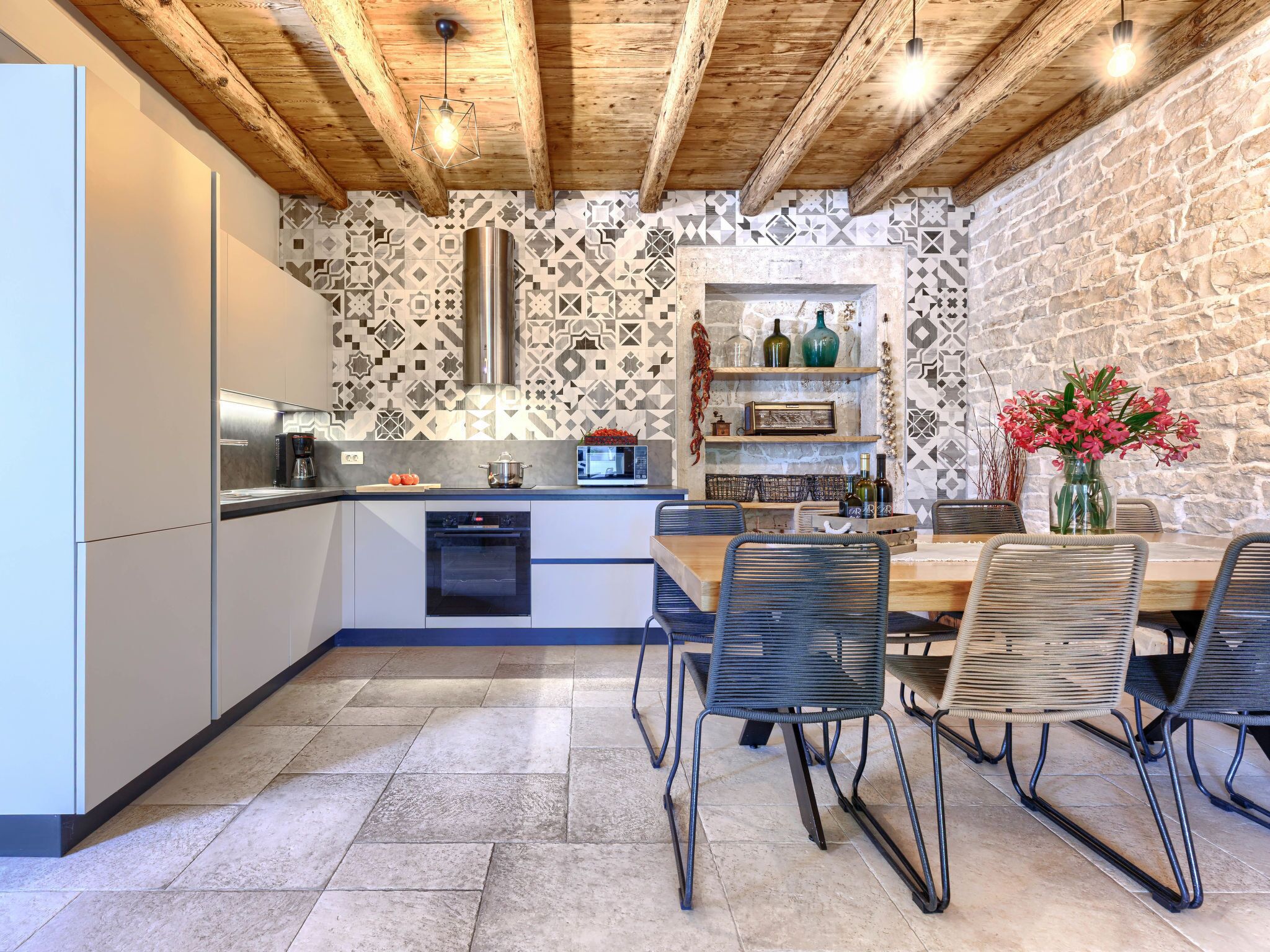 Una cocina moderna con salpicadero de piedra, vigas de techo de madera y un comedor con sillas de metal y una mesa de madera.