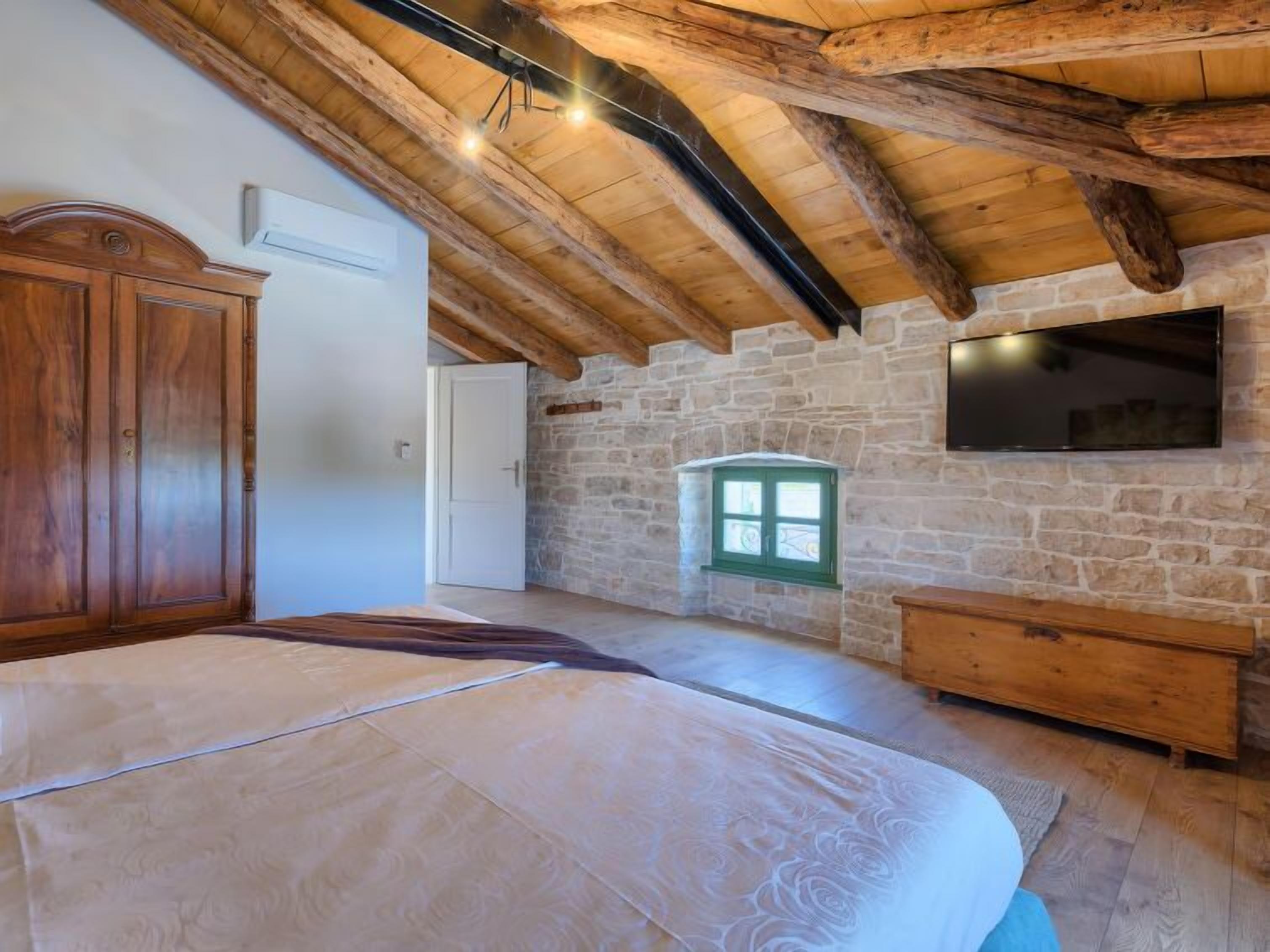 A bedroom with a wooden ceiling, stone walls, a bed, a wardrobe, a TV, and a small window.