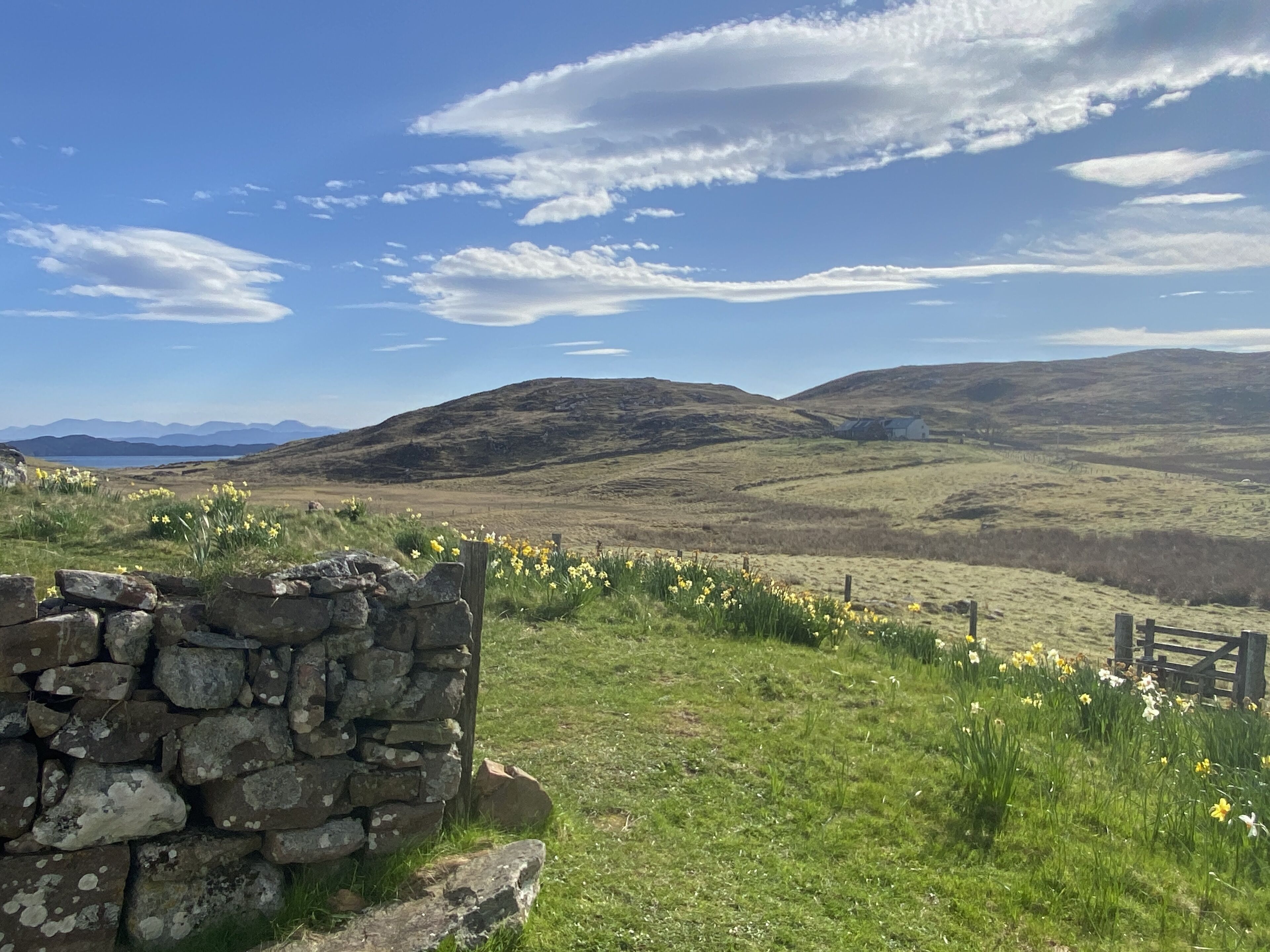 A stone wall with a grassy field and yellow flowers in the foreground, a stone building in the middle ground, and rolling hills in the background under a clear sky.