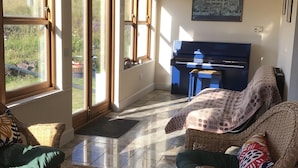 Ein geräumiges Zimmer mit Korbmöbeln, einem Klavier und großen Fenstern mit Blick ins Freie.