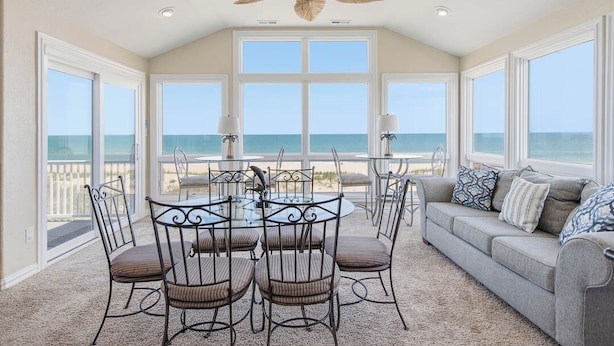 A room with a large window overlooking the ocean, a ceiling fan, a round dining table with chairs, and a sofa with cushions. - Quiet Time | Oceanfront (Virginia Beach)