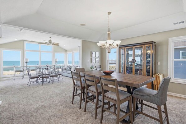 A spacious dining area with a large wooden table and chairs, a chandelier, and a view of the ocean through large windows.