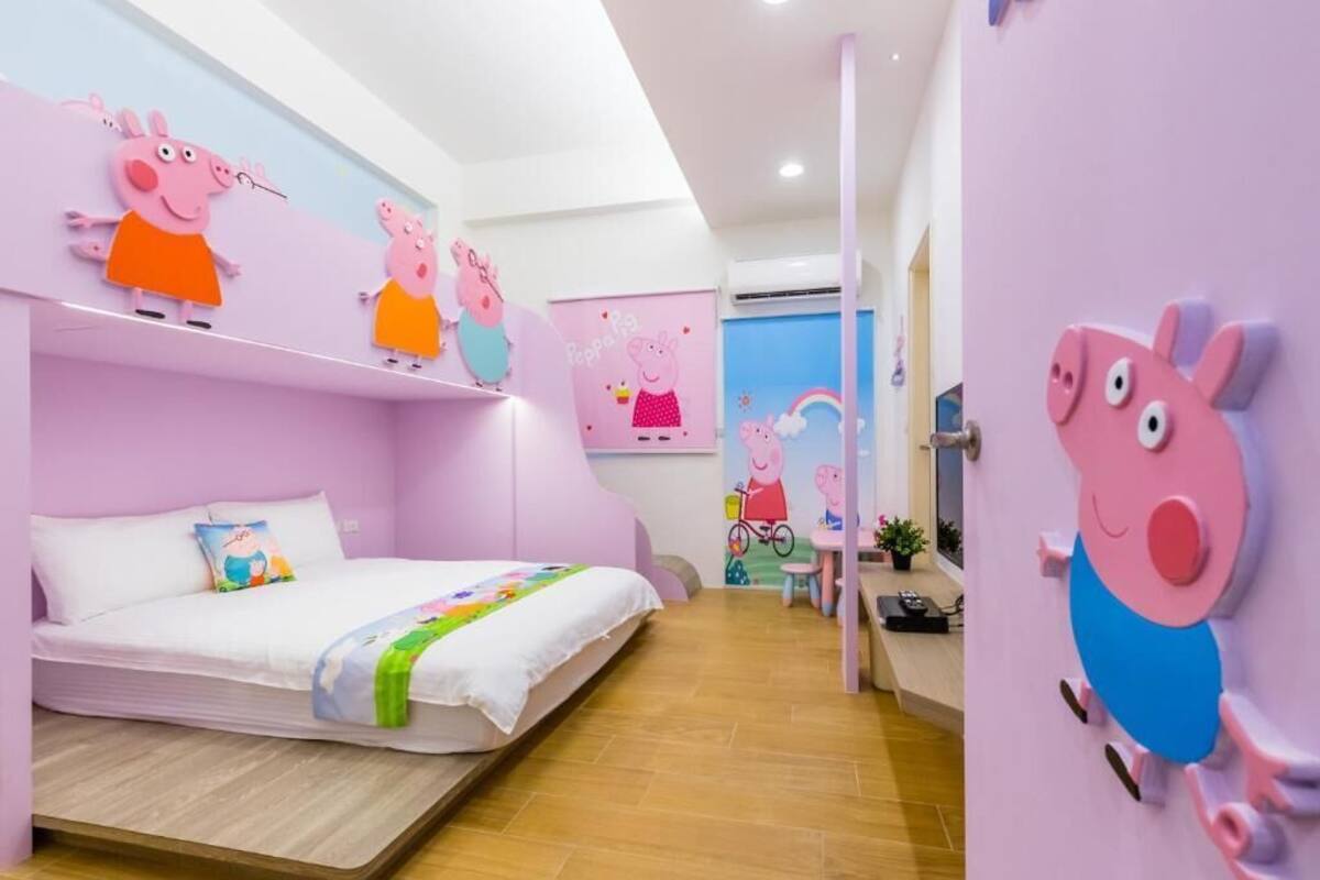 A children's room with a pink theme, featuring wall decorations of a cartoon pig character, a bed with a colorful blanket, and a small table with a chair.