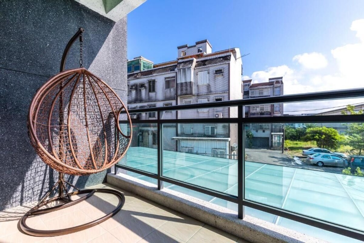 A balcony with a wicker chair, a glass railing, and a view of a parking lot and buildings.