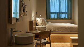 A modern bedroom with a bed, bedside table, lamp, and a view of the outdoors through a window.
