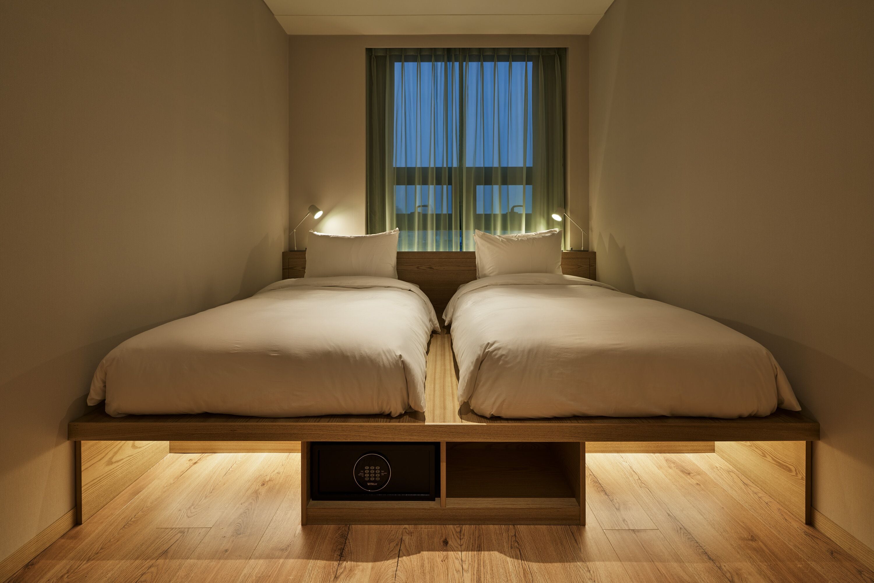 A bedroom with two beds, a window with curtains, bedside lamps, and a built-in storage compartment.