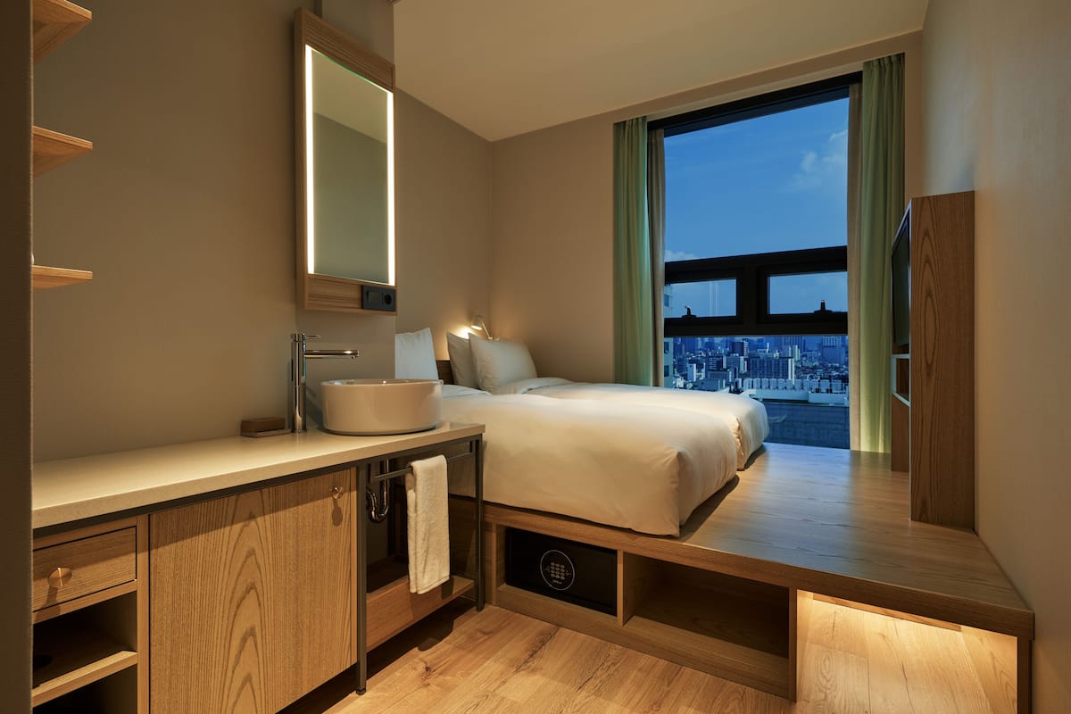 A modern hotel room with a large bed, a vanity with a mirror, a sink, and a view of the cityscape through a large window.