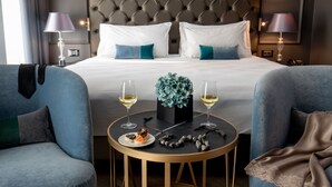 Habitación de hotel con una cama grande, dos lámparas de noche, una mesa redonda con una planta y un plato de comida, y dos sillones.
