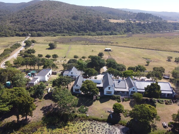 An aerial view of a rural property with multiple buildings, a dirt road, and surrounding fields. - Bitou River Lodge (Plettenberg Bay)