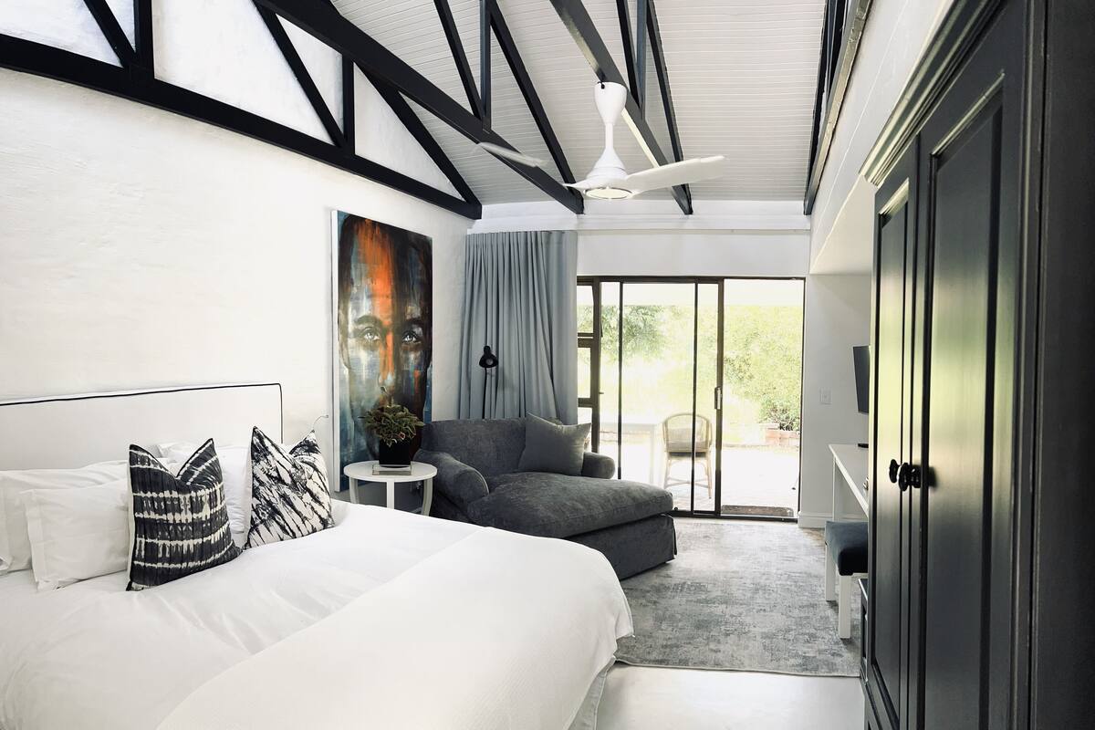 Quarto moderno com uma cama grande, um sofá cinza, um ventilador de teto e uma porta de vidro deslizante que dá para uma área externa.