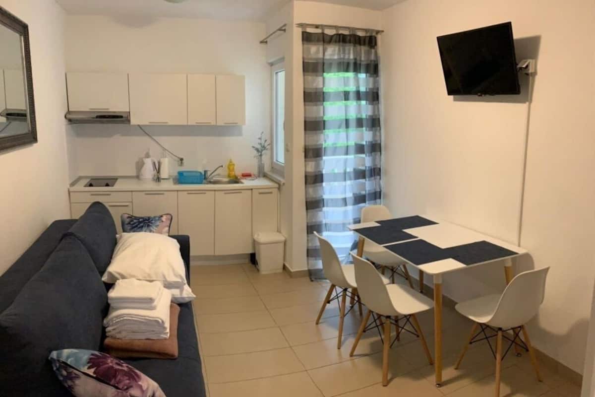 A compact living space with a sofa, a dining table with chairs, a small kitchenette, and a TV mounted on the wall.