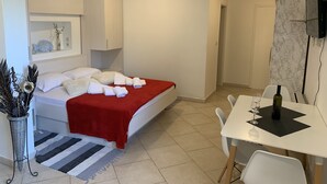 Una habitación de hotel con cama, una mesa de comedor con sillas, un televisor montado en la pared y una pequeña planta en maceta.