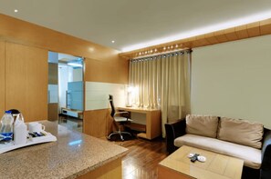 A modern hotel room with a wooden desk, a sofa, a coffee table, and a bathroom visible in the background. - Hotel Balaji Central (Ratlam)