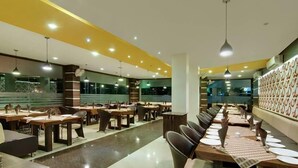 Een ruime restaurantruimte met meerdere eettafels en stoelen, een centrale doorgang en een patroonwand aan de ene kant.
