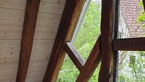 A room with a wooden ceiling, a window with a view of greenery, and a small round table with a chair.