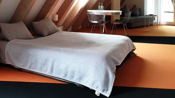 A bedroom with a sloped ceiling, a bed, a small table with chairs, and a sofa.