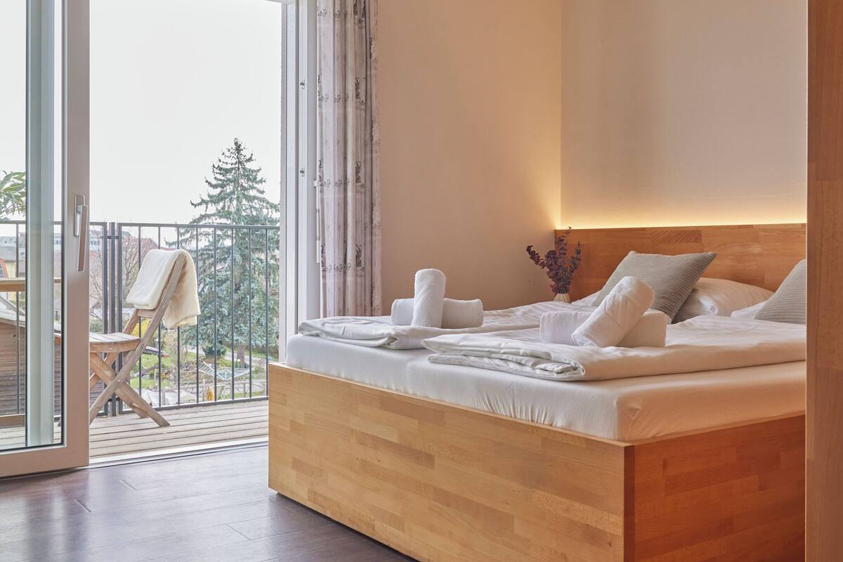 Une chambre moderne avec un lit en bois, une porte en verre donnant sur un balcon et une vue sur les arbres.