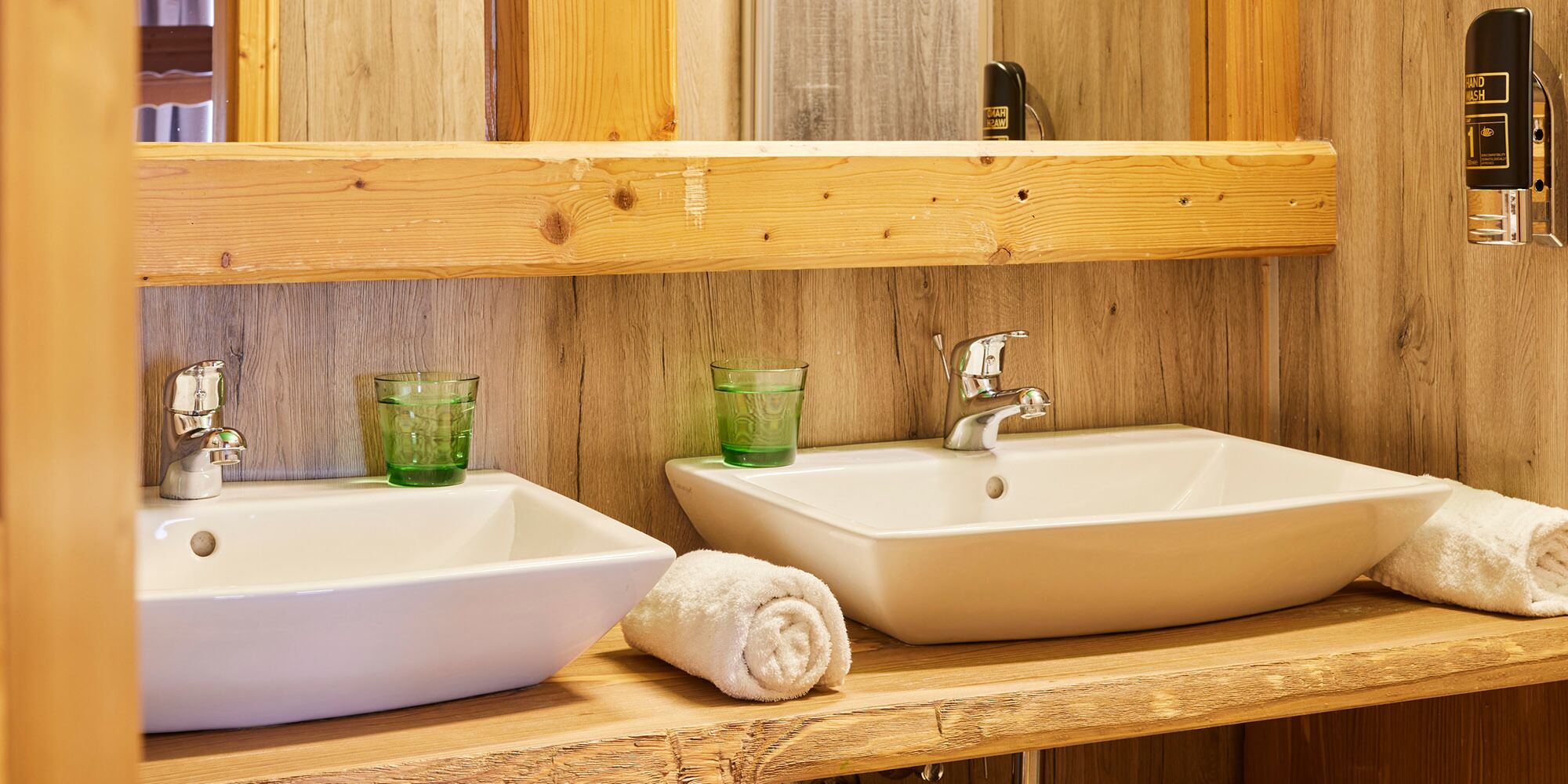 A bathroom with two white sinks, green glass cups, a towel, and a wooden wall.