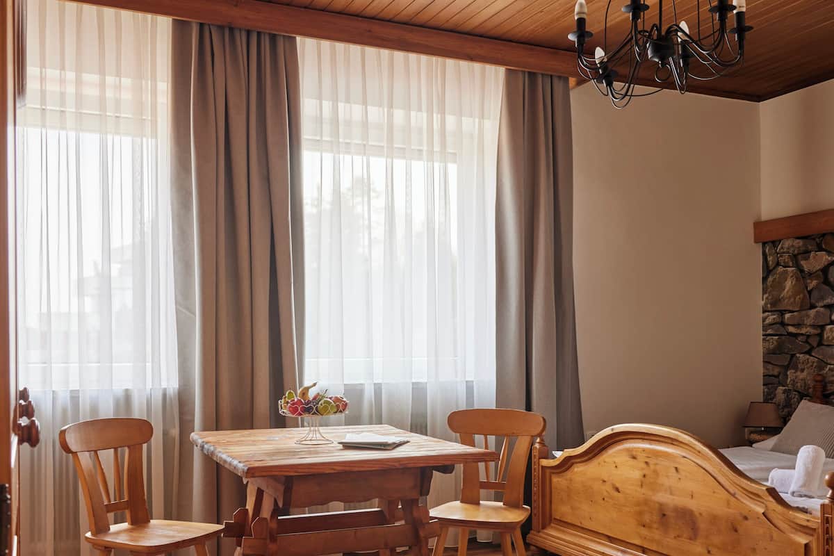 Une pièce avec un plafond en bois, un lit doté d’une tête de lit en pierre, une table en bois avec des chaises et une fenêtre avec des rideaux.