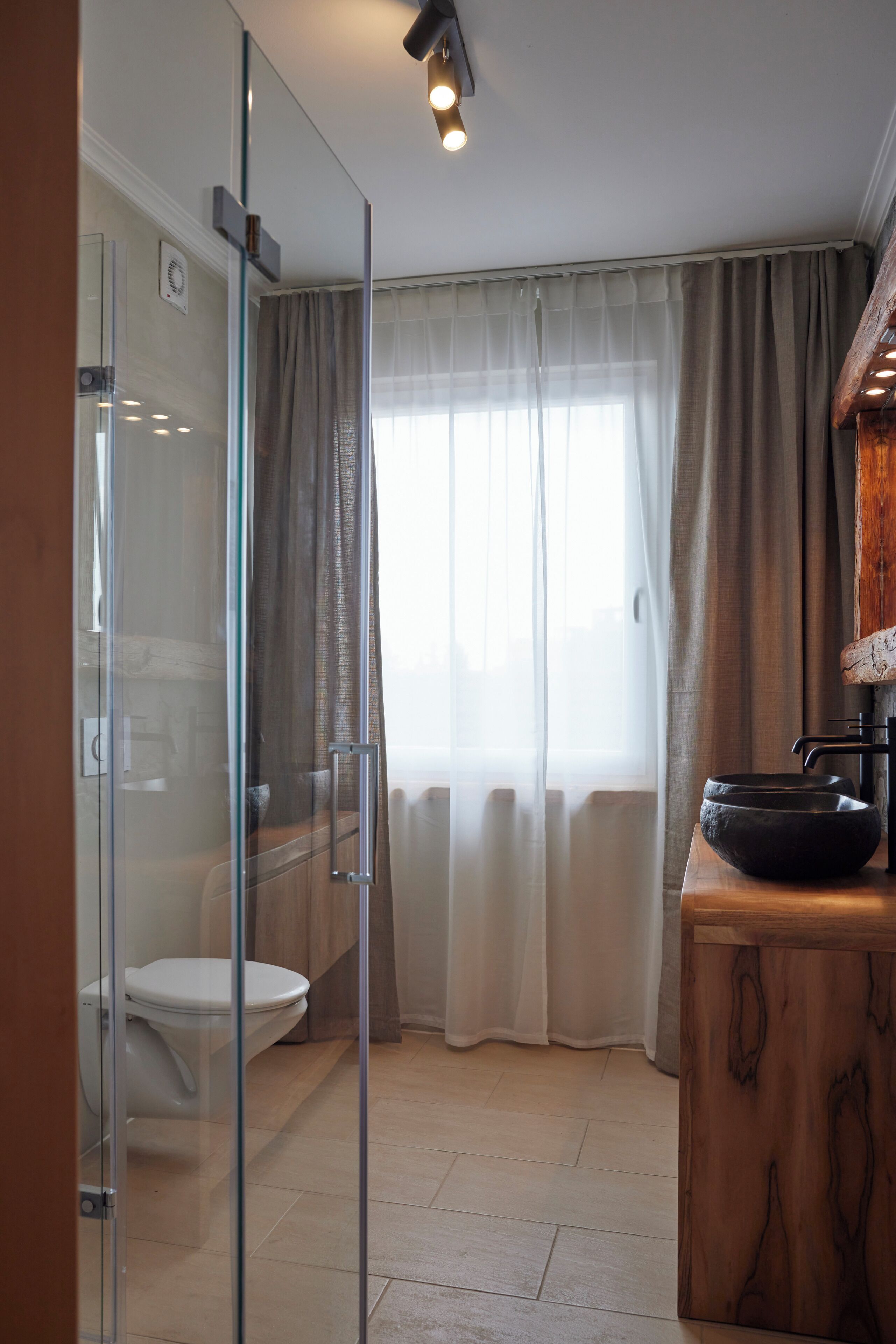 Un baño moderno con cabina de ducha de vidrio, inodoro blanco, tocador de madera con lavabo negro y un ventanal con cortinas translúcidas.