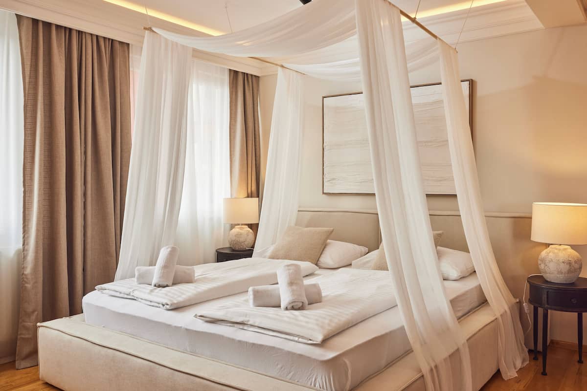 Un lit à baldaquin avec du linge de lit et des serviettes blanches, une table de chevet avec une lampe, et une grande œuvre d’art encadrée au mur.
