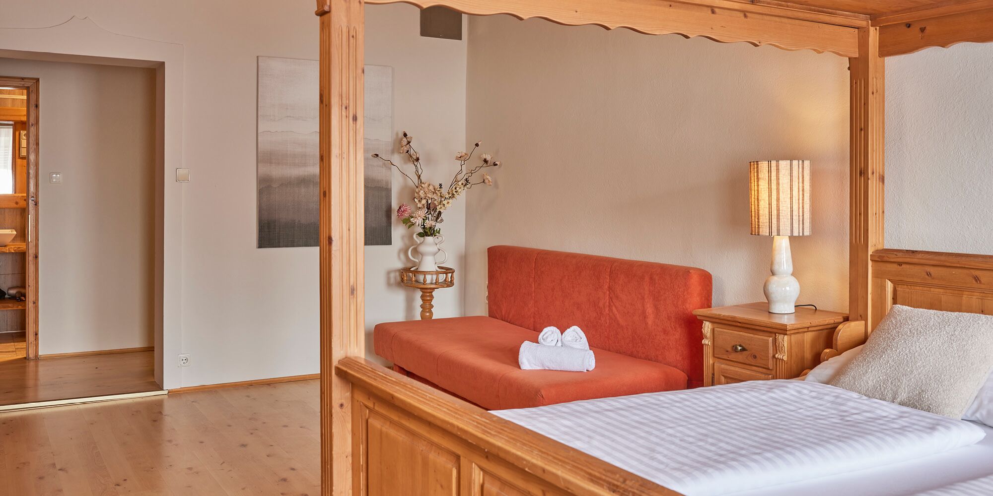Un dormitorio con cama con dosel, una mesita de noche con lámpara, un jarrón con flores y un cuadro en la pared.