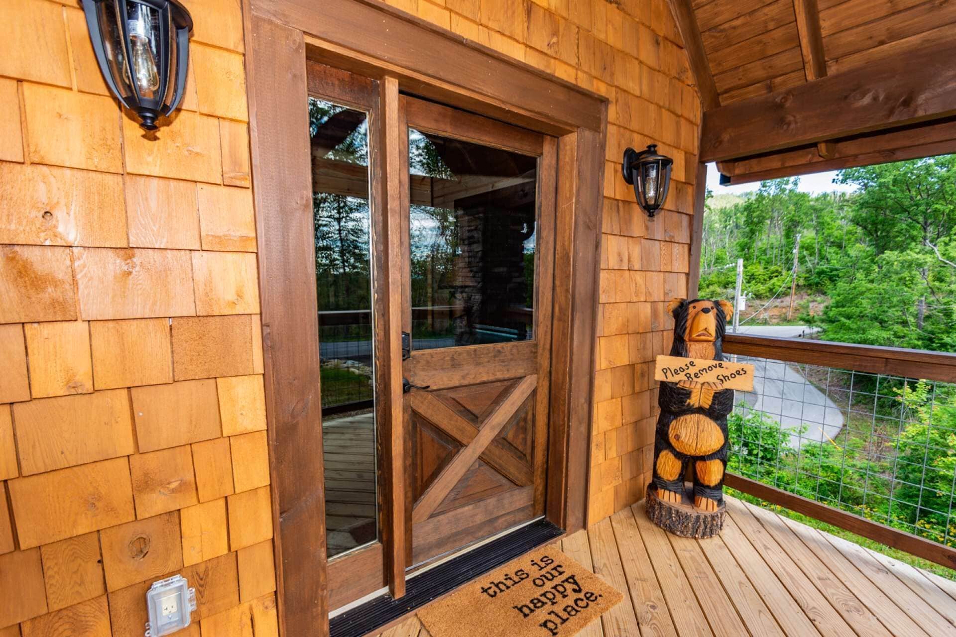 A wooden cabin entrance with a glass door, a bear sculpture holding a sign, and a doormat that reads "this is our happy place."