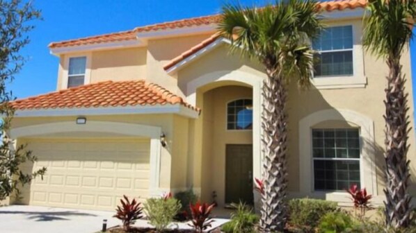 A two-story house with a red-tiled roof, a garage, and a well-maintained front yard with palm trees and red-leafed plants. - Dream Vacation Home With CDC Cleaning Standards! Stunning and Spacioud 6BD Villa Near Disney! #6av350 (Davenport)