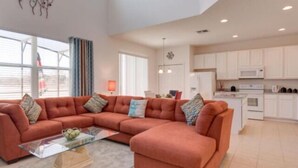 A modern living room with a sectional sofa, a glass coffee table, and a kitchen area with white cabinets and appliances. - Dream Vacation Home With CDC Cleaning Standards! Stunning and Spacioud 6BD Villa Near Disney! #6av350 (Davenport)
