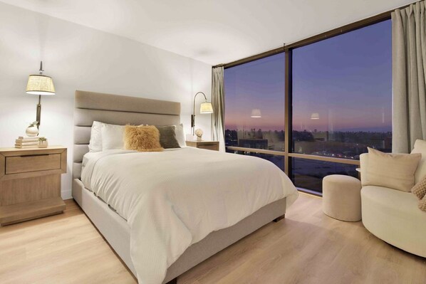 A modern bedroom with a large bed, a nightstand, a lamp, a chair, and a large window with a city view. - Luxury Smart Home w/Gorgeous Waterfront Retreat + Amazing Sunset Views (Marina Del Rey)