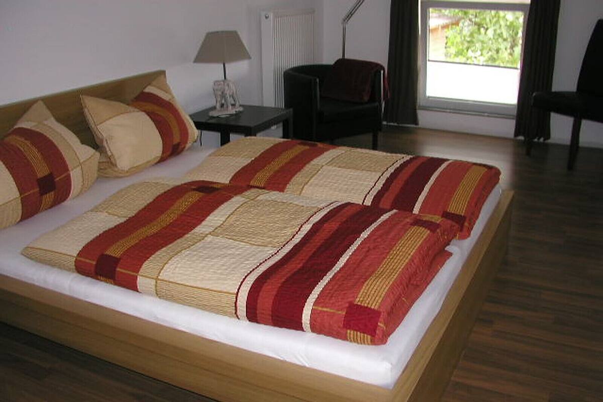 A neatly made bed with a striped blanket and pillows, a small bedside table with a lamp, a chair, and a window with a view of greenery.