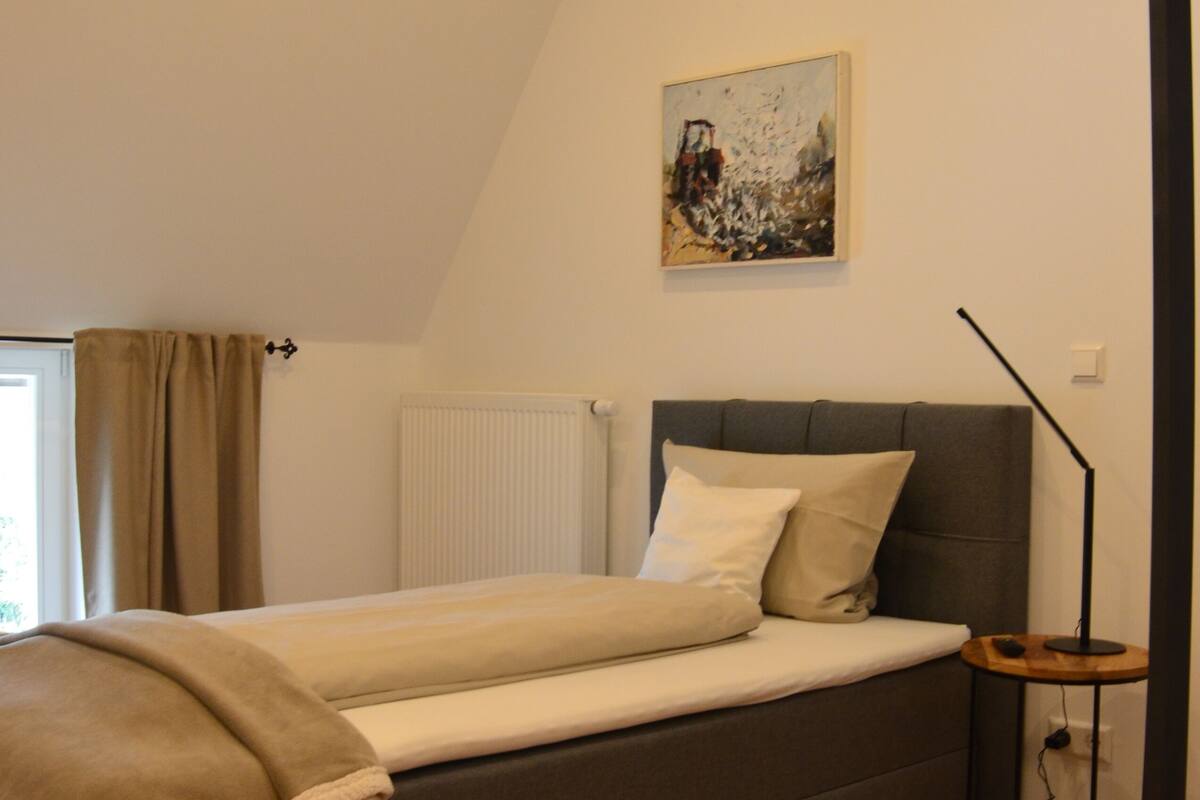 A bedroom with a bed, a radiator, a painting on the wall, and a floor lamp.