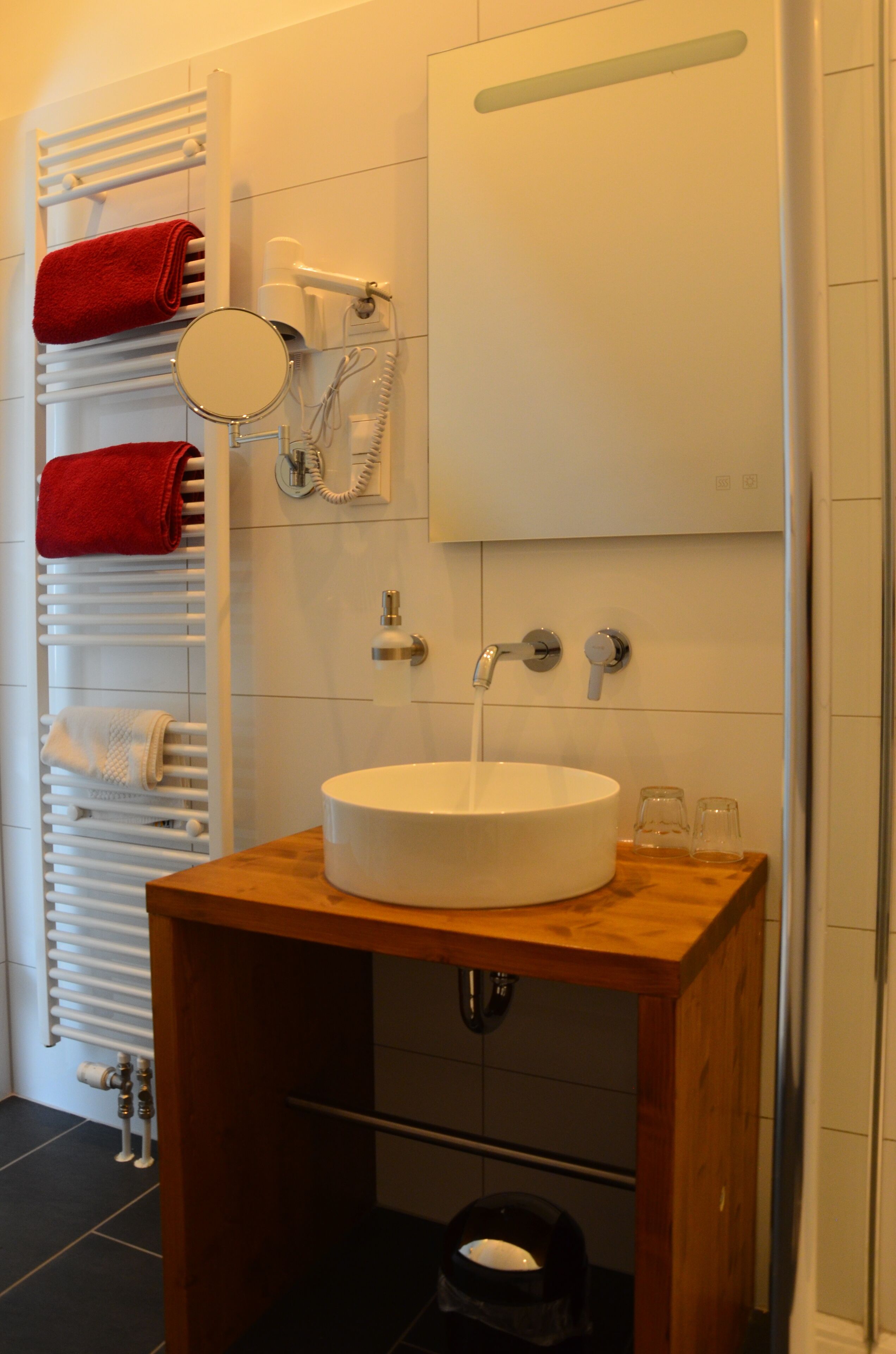 Un baño con tocador de madera, lavamanos blanco, espejo, toallero con toallas rojas y zona de ducha.