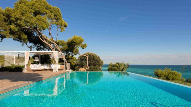 Una piscina infinita de color azul intenso con un área de estar exterior blanca y una pérgola, con vistas al mar.