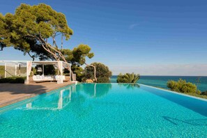 En vacker, turkosblå infinitypool med ett vitt utomhusområde med sittplatser och en pergola, med utsikt över havet.