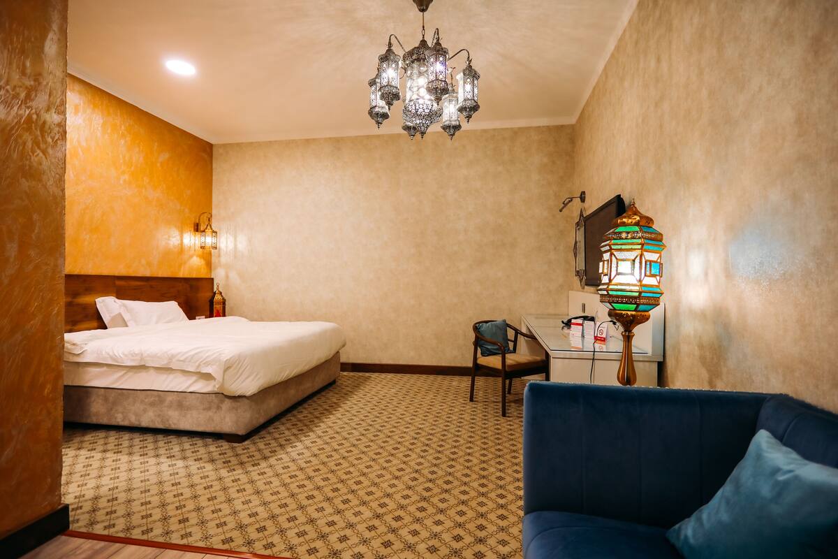 A hotel room with a bed, a blue sofa, a desk with a lamp, and a chandelier.