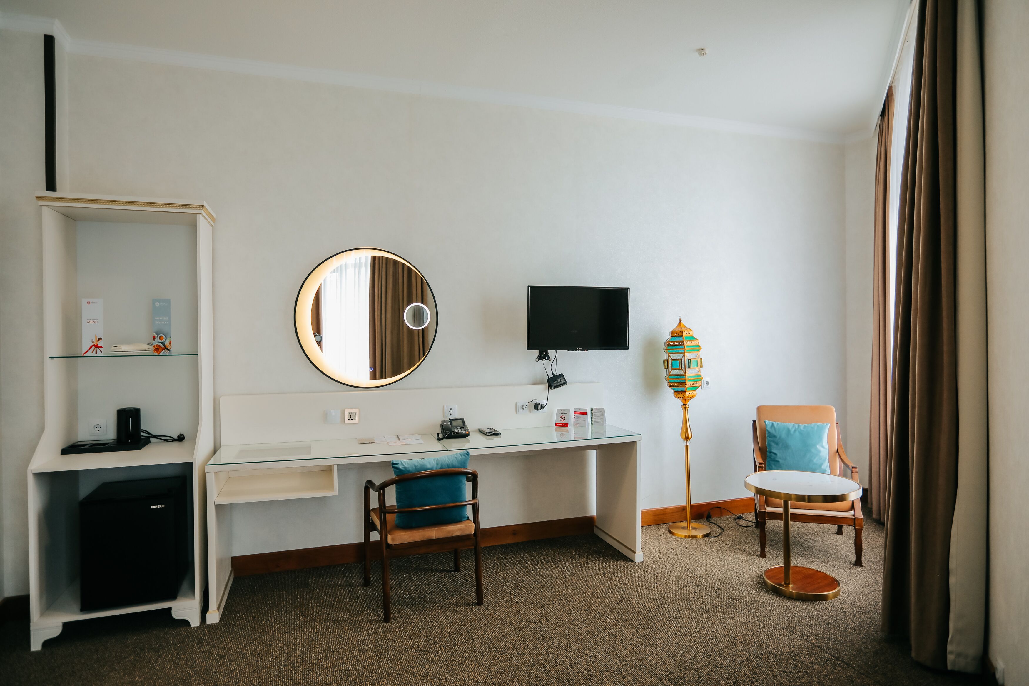 A hotel room with a desk, a chair, a television, a bookshelf, and a floor lamp.