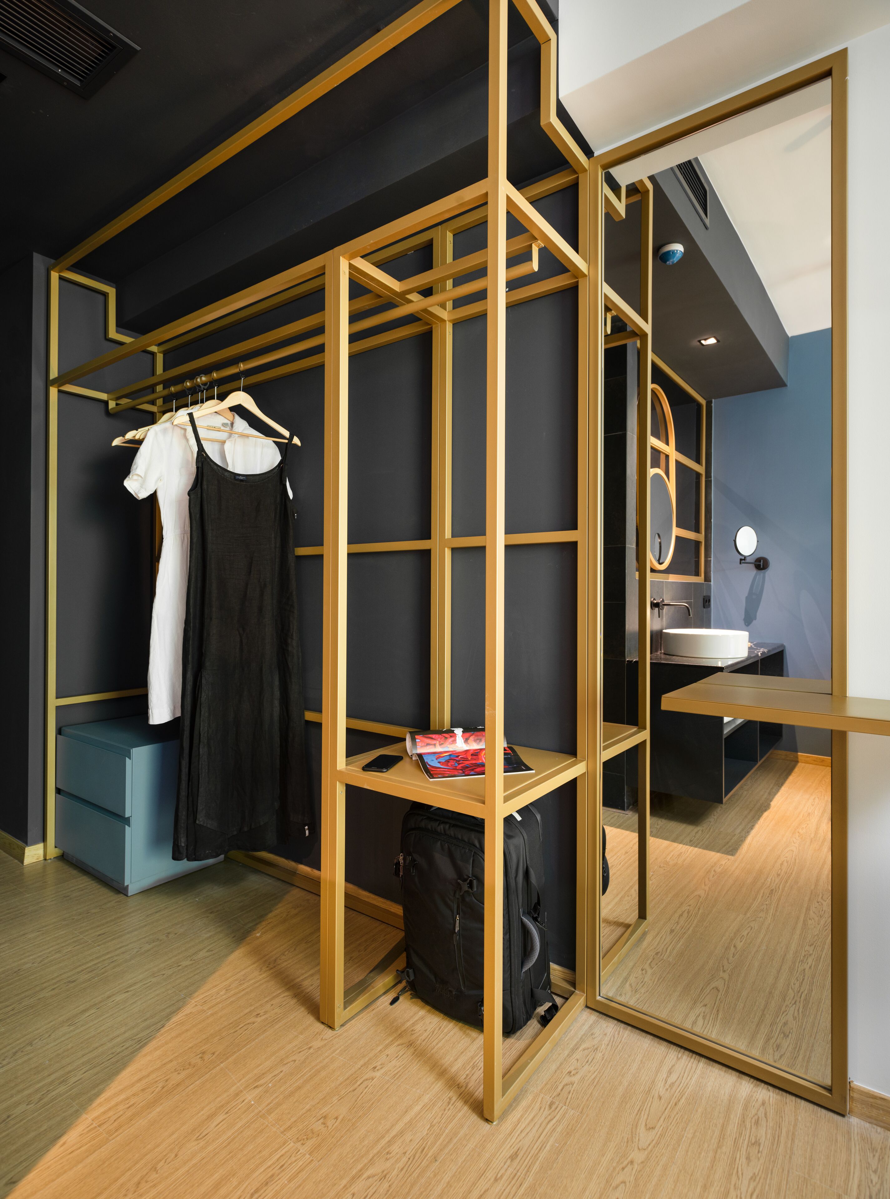Une chambre moderne avec un séparateur en bois, une table de chevet bleue, un sac noir et une armoire suspendue contenant des vêtements.