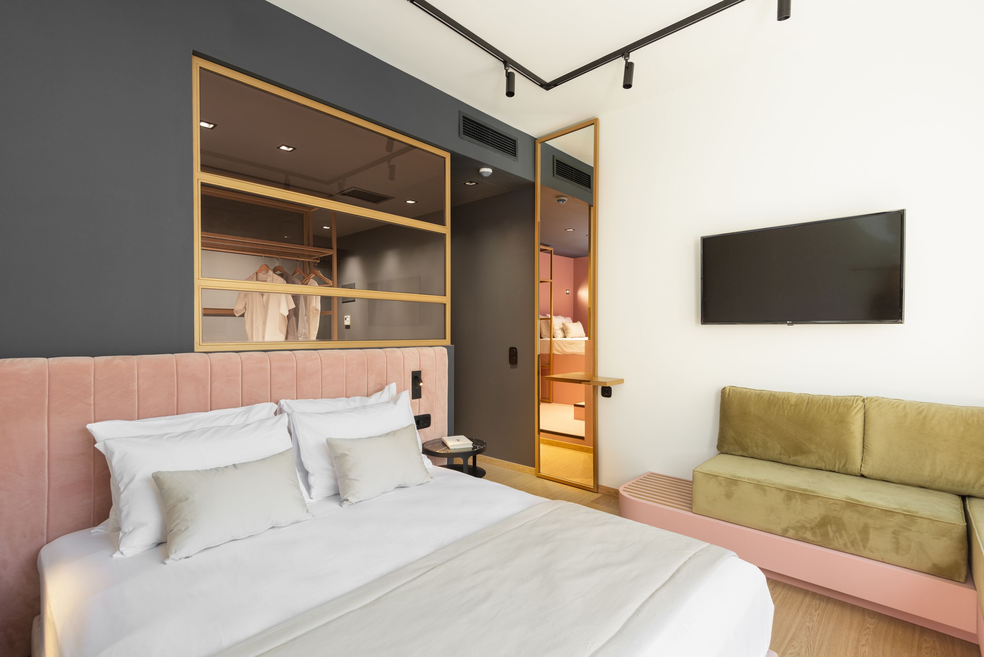 A modern hotel room with a large bed, a seating area, a wardrobe with a mirror, and a flat-screen TV.