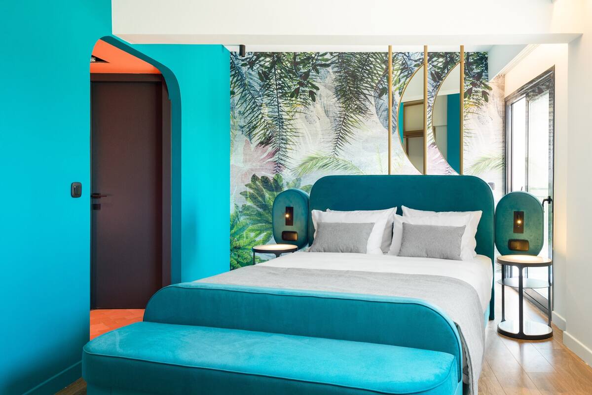 A modern bedroom with a teal color scheme, a large bed with a matching headboard, two bedside tables with lamps, and a large window with a view of palm trees.
