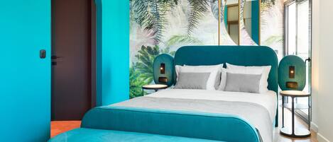 A modern bedroom with a teal color scheme, a large bed with a matching headboard, two bedside tables with lamps, and a large window with a view of palm trees.