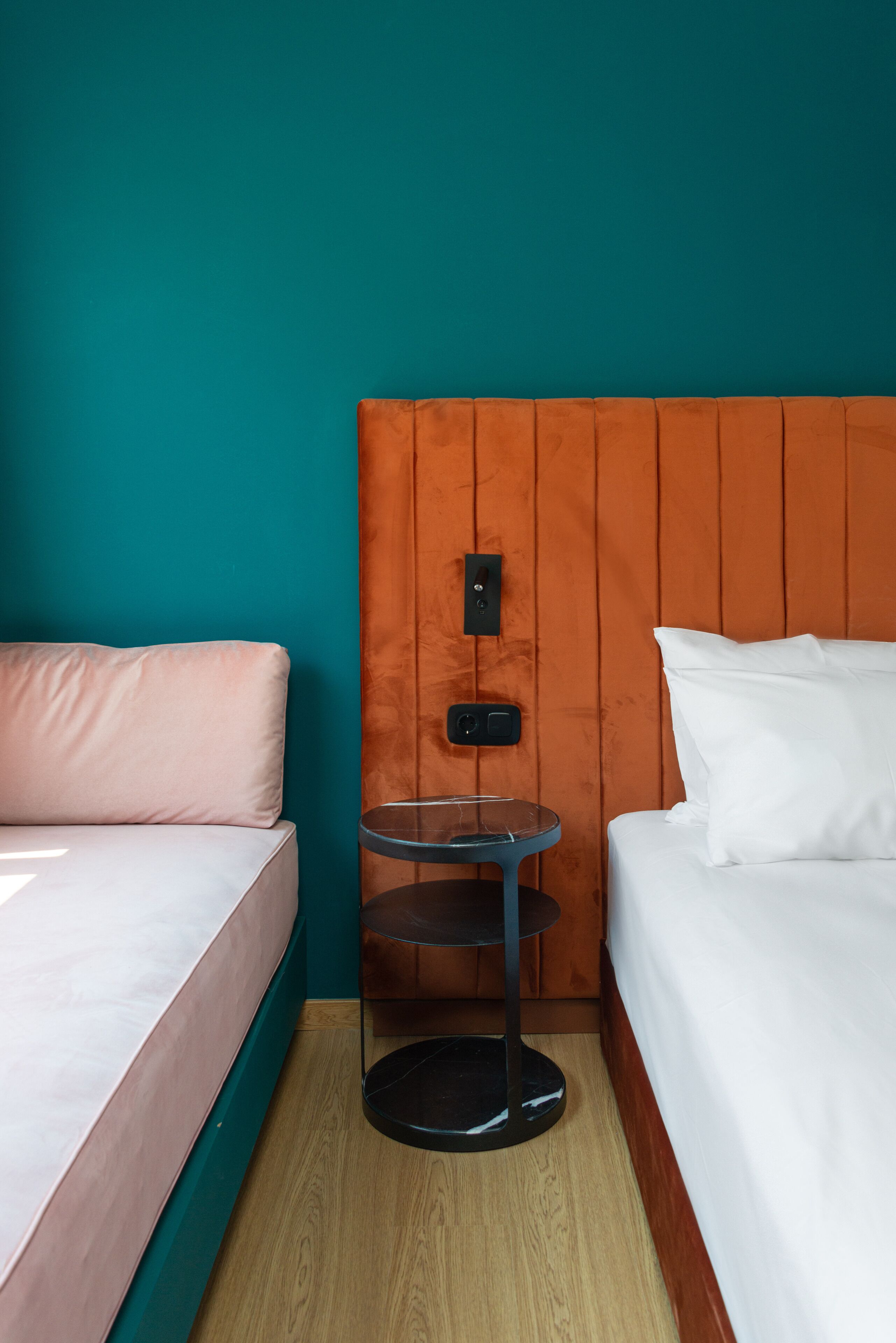 A hotel room with teal walls, a wooden headboard, a bed with white bedding, a pink sofa, and a small round table with a black base.