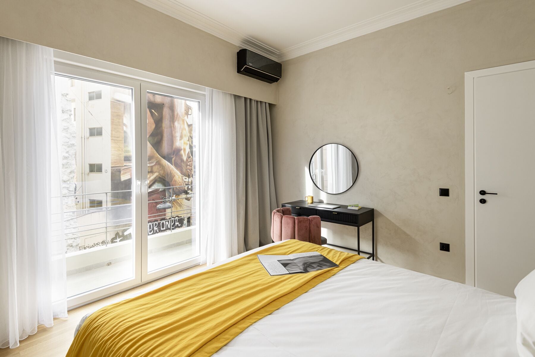 Ein Hotelzimmer mit Bett, Nachttisch mit Spiegel, Fenster mit Vorhängen und einer Tür.