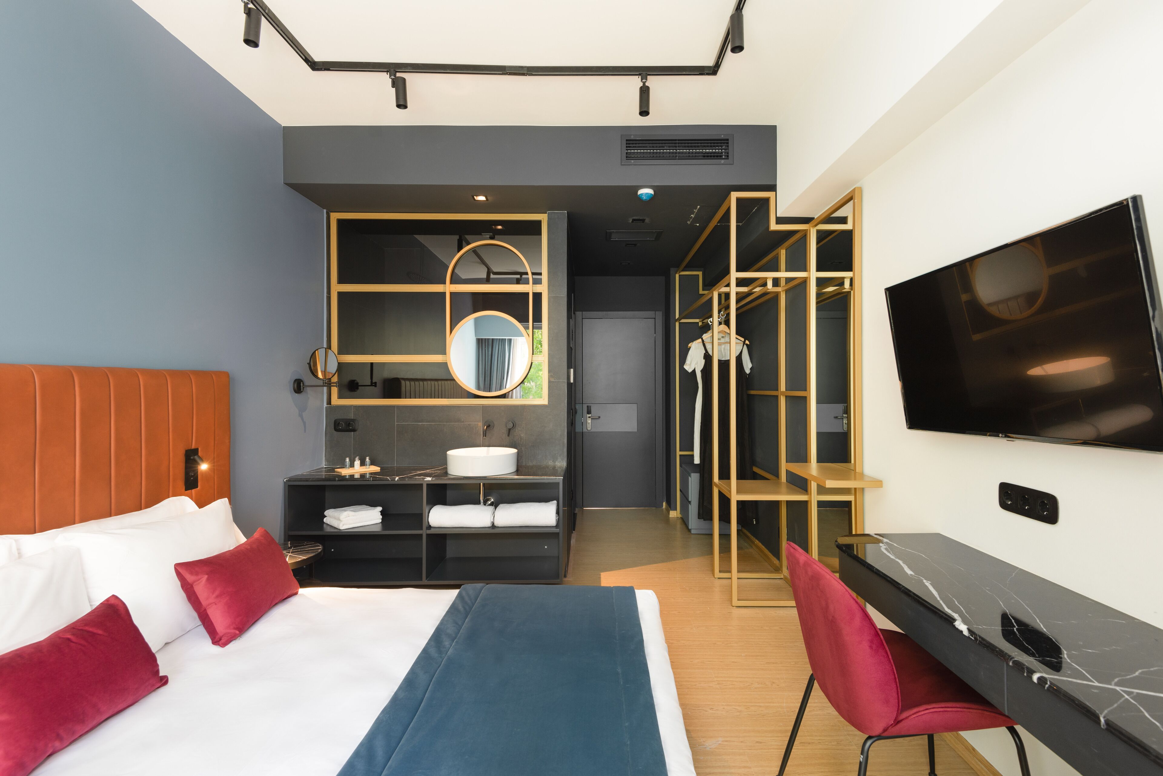 A modern hotel room with a bed, a desk, a TV, a bathroom with a mirror, and a wardrobe.