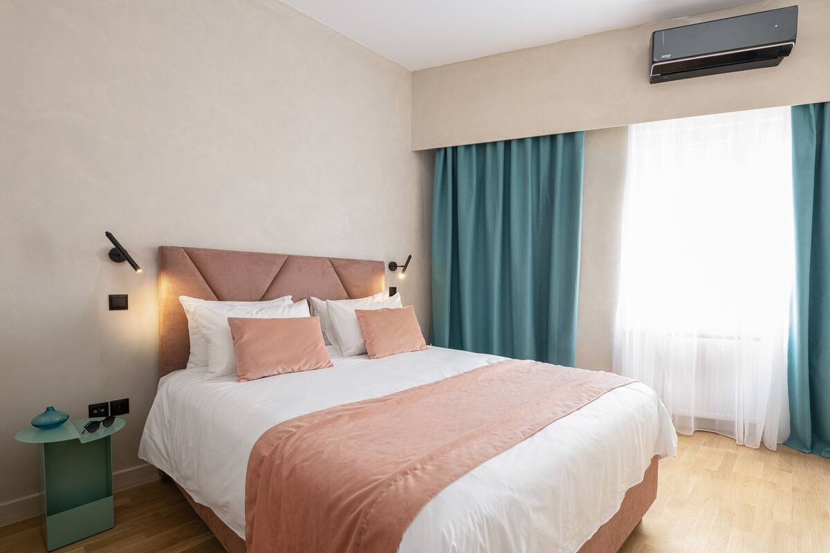 A modern hotel room with a large bed, a bedside table with a lamp, and a window with curtains.