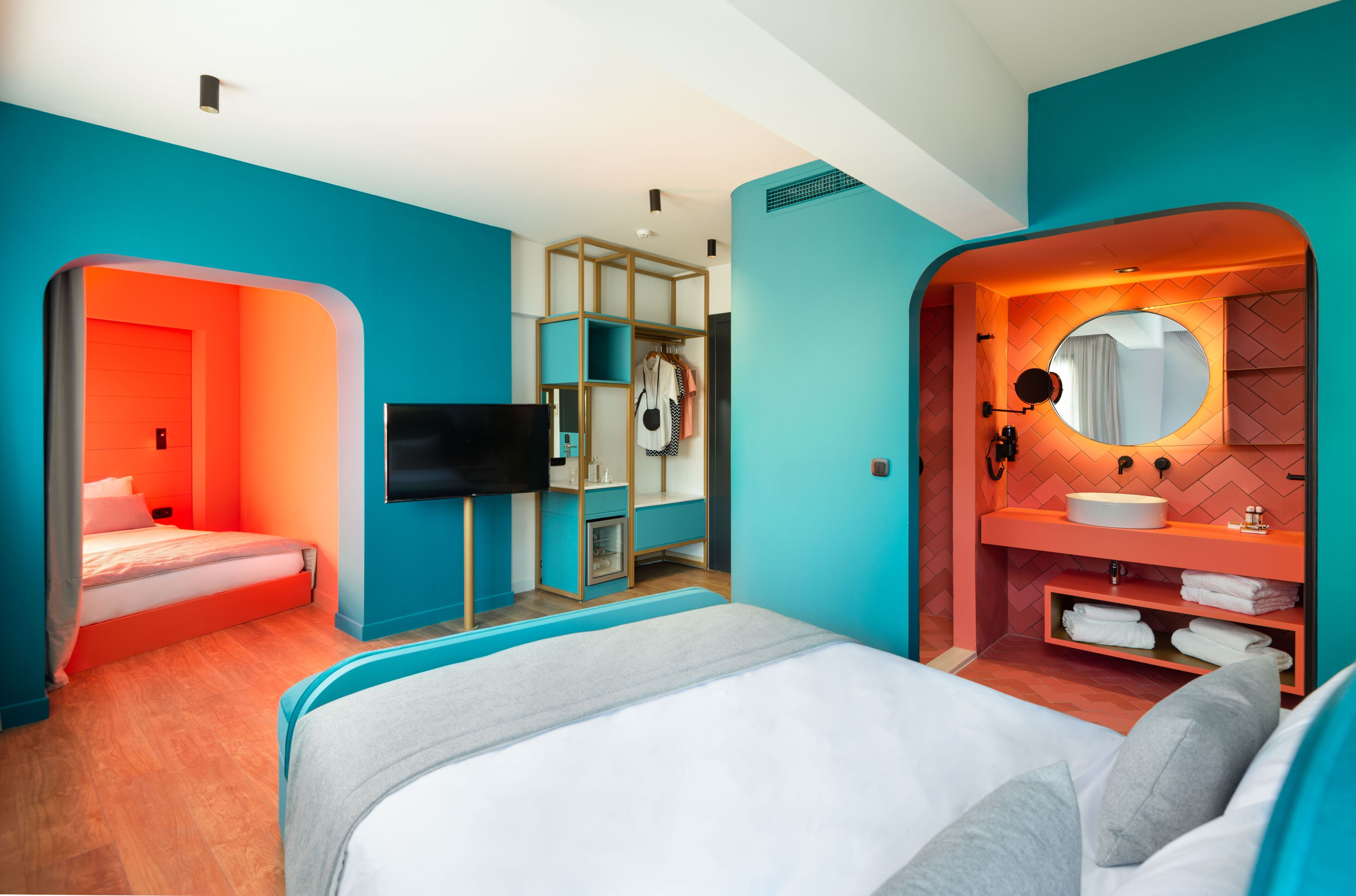 Ein modernes Hotelzimmer mit einem großen Bett, einem Fernseher, einem begehbaren Kleiderschrank und einem Badezimmer mit rundem Spiegel und Waschbecken.