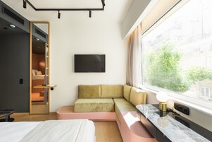 Quarto de hotel moderno com cama, área de estar com sofá e banquinho, TV fixada na parede e janela com vista para edifícios e árvores.