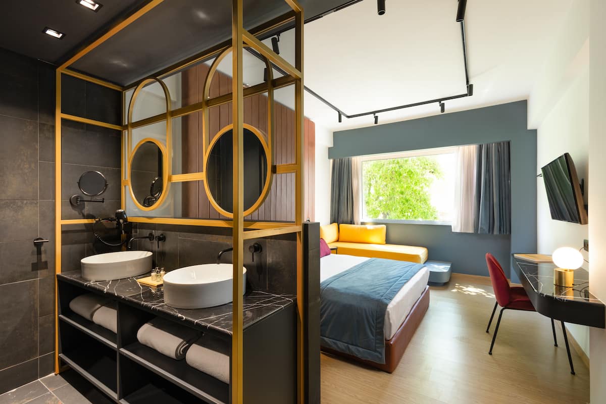 A modern hotel room with a dual sink vanity, a bed with a blue bedspread, a desk with a lamp, and a large window with curtains.